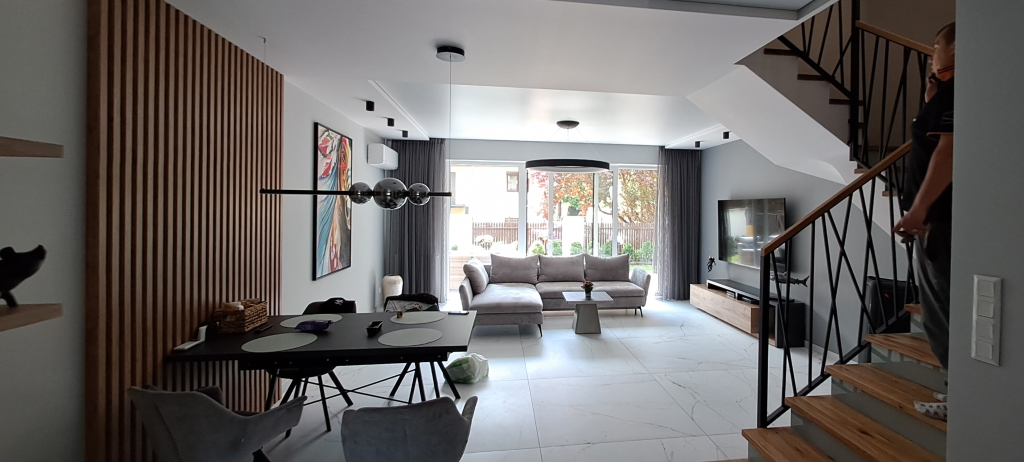 Nowoczesny salon z drewnianą ścianą z lameli, szarą sofą narożną, marmurową podłogą i czarnymi schodami z drewnianymi stopniami.