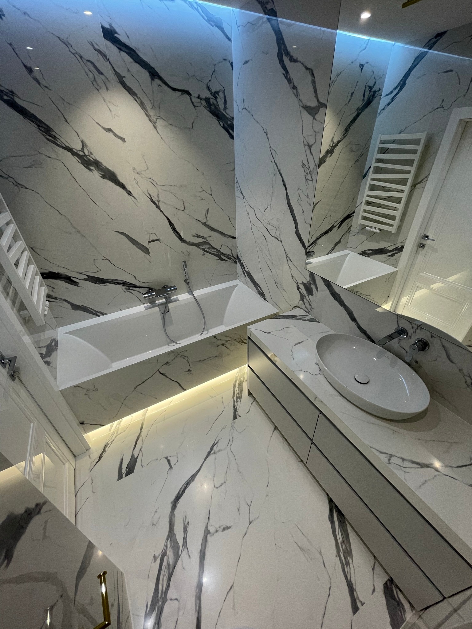 Nowoczesna łazienka wykończona białym marmurem z czarnymi żyłami, oświetlenie LED pod wanną i szafką umywalkową, owalna umywalka nablatowa.