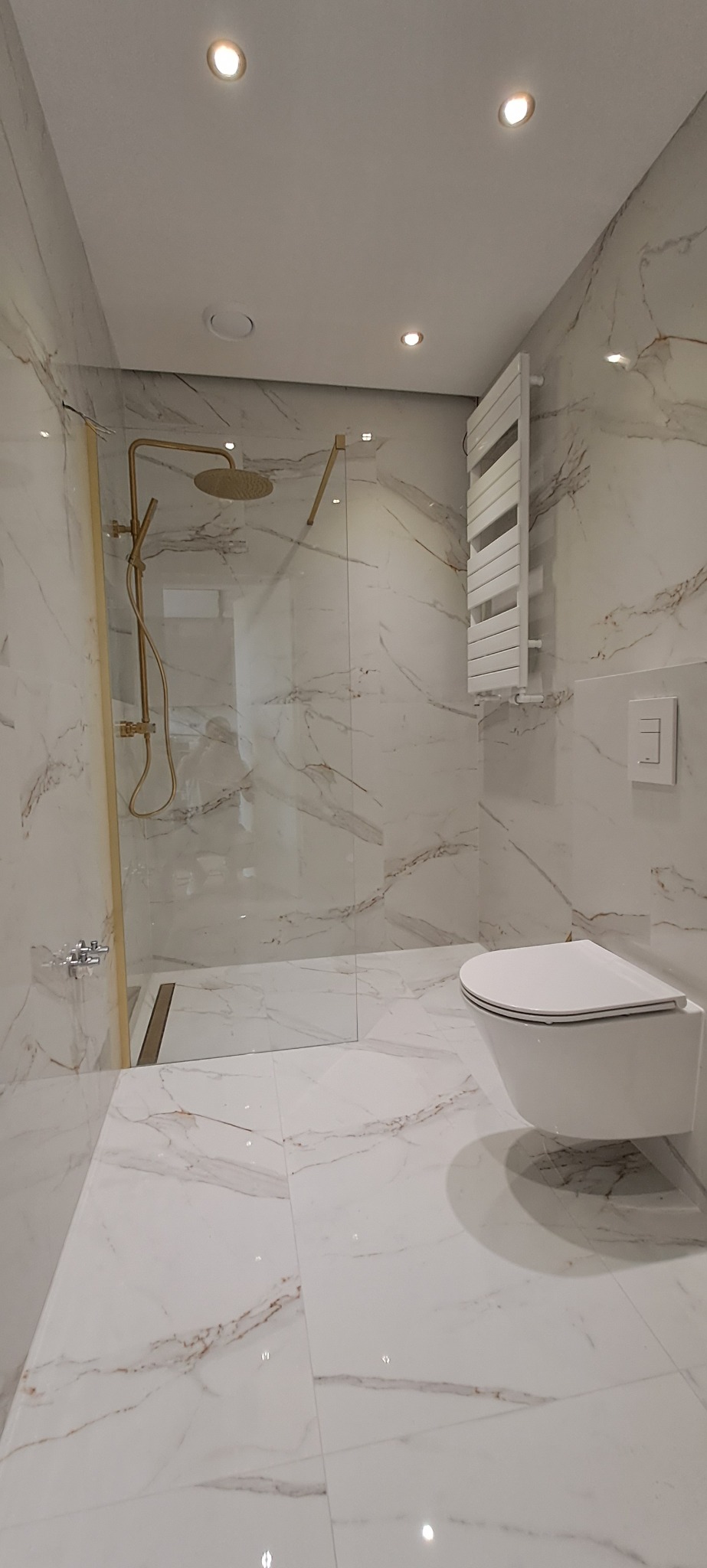 Nowoczesna łazienka z kabiną prysznicową typu walk-in z armaturą w kolorze złota, wiszącą miską WC, grzejnikiem drabinkowym i białymi płytkami imitującymi marmur.