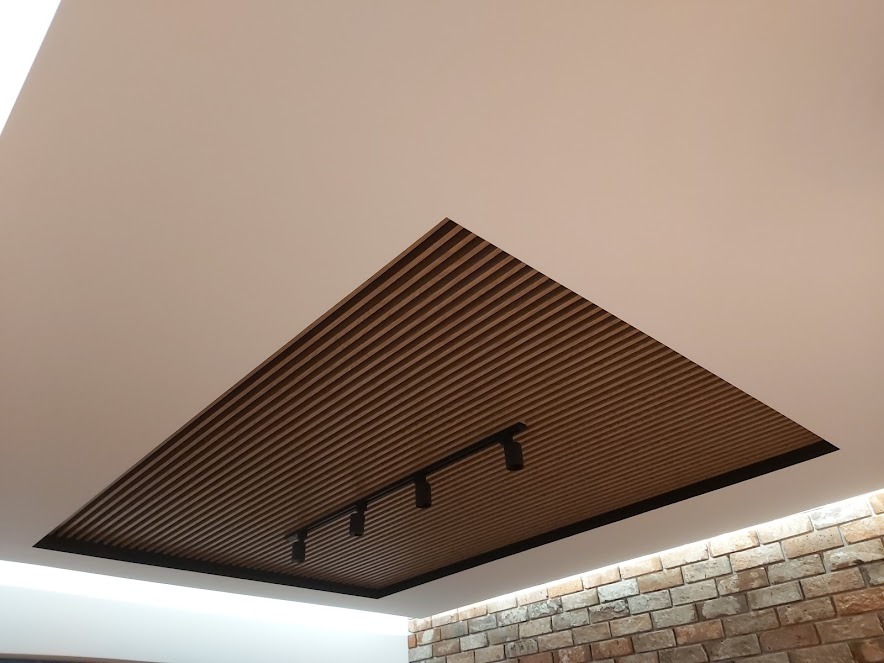 Nowoczesny sufit podwieszany z drewnianymi lamelami i czarnym oświetleniem punktowym, widoczny fragment ściany z cegły.