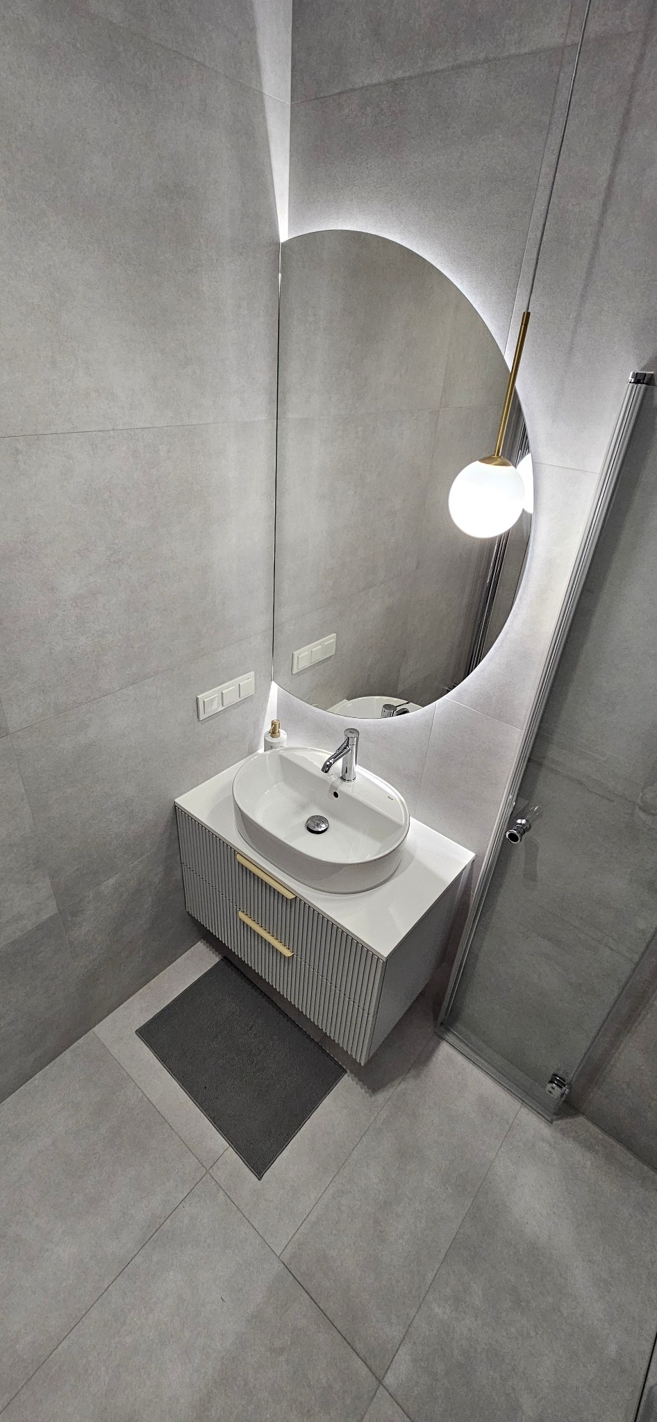 Nowoczesna łazienka w odcieniach szarości z owalną umywalką nablatową, szafką z ryflowanymi frontami i okrągłym lustrem podświetlanym LED, uzupełniona o wiszącą lampę z kulistym kloszem i szklaną...