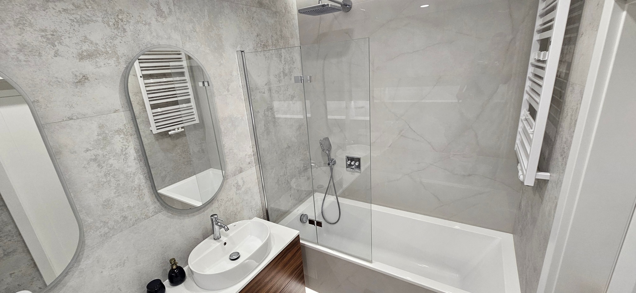 Nowoczesna łazienka z owalnym lustrem, szklanym parawanem nawannowym, białą wanną i grzejnikiem drabinkowym na tle szarych płytek imitujących kamień.