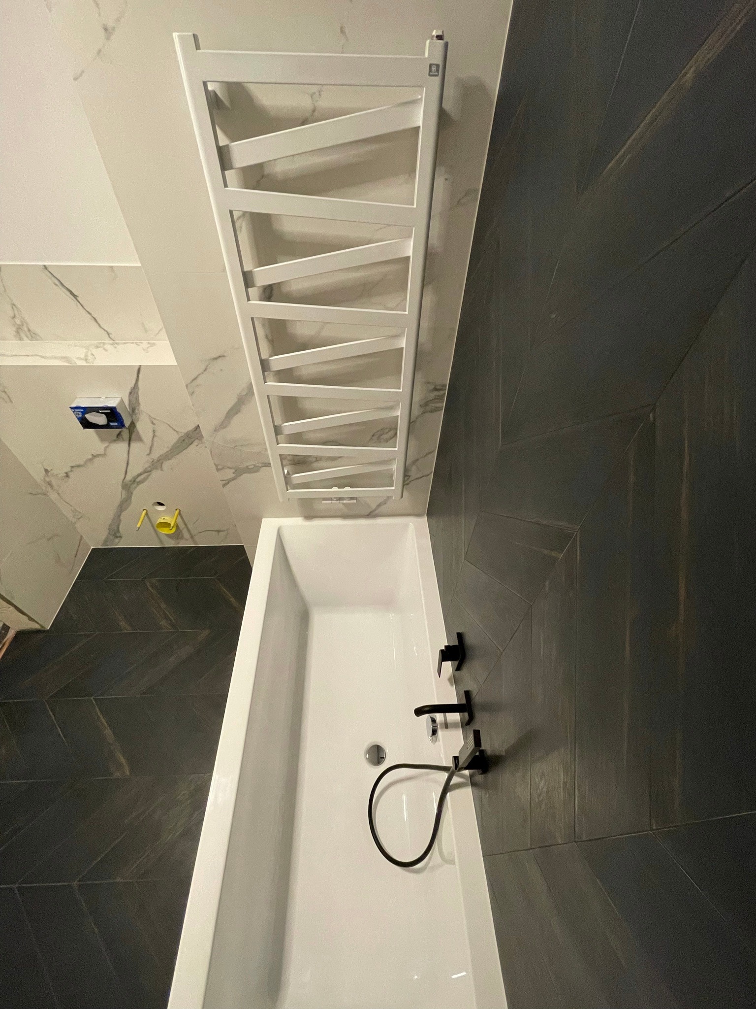 Wanna z czarną armaturą na tle ciemnych płytek ułożonych w jodełkę, biały grzejnik drabinkowy, fragment ściany z marmurowymi płytkami.