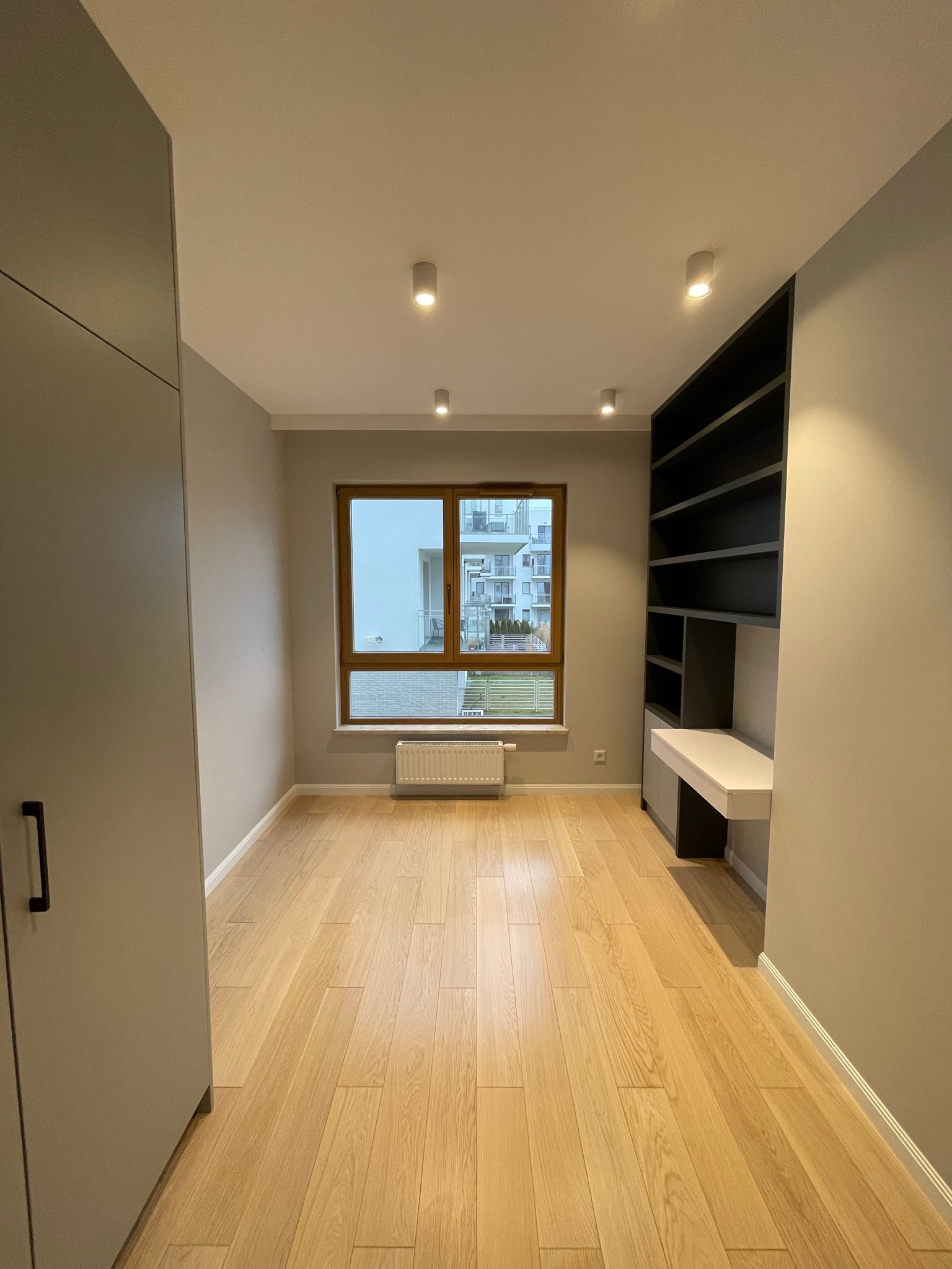 Pokój po remoncie: jasna drewniana podłoga, szara szafa w zabudowie, ciemne regały z biurkiem, okno z widokiem na budynek.