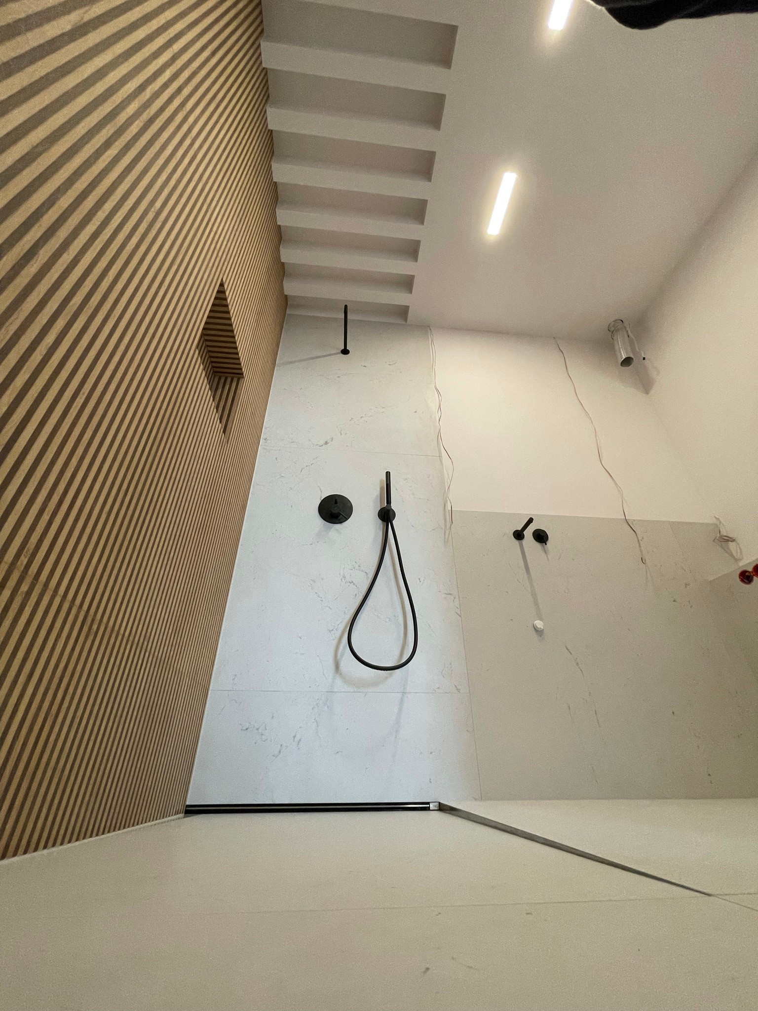 Nowoczesna kabina prysznicowa walk-in z drewnianą ścianą z lamelami, czarną armaturą i białymi płytkami imitującymi marmur, oświetlenie LED w suficie.