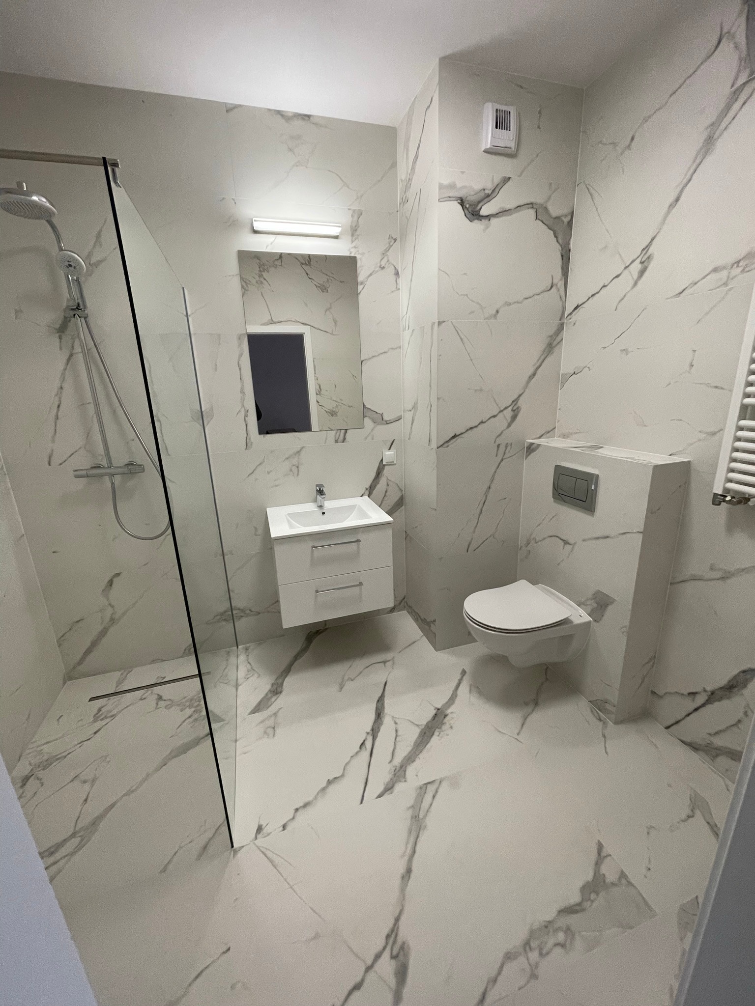 Nowoczesna łazienka wykończona białymi płytkami imitującymi marmur, z kabiną prysznicową typu walk-in, wiszącą szafką z umywalką i podwieszaną toaletą z zabudowanym stelażem.