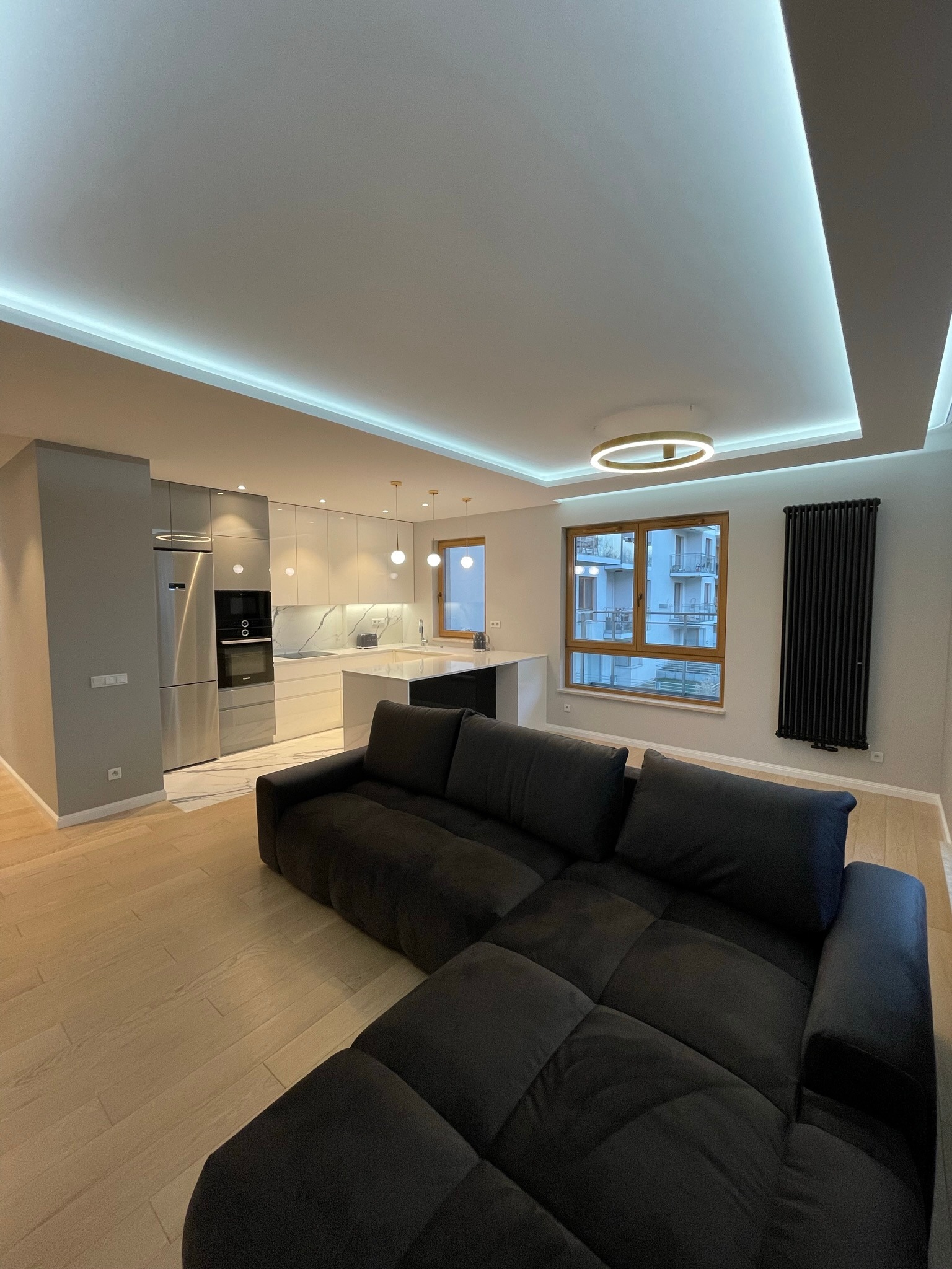 Nowoczesne wnętrze mieszkania z otwartą kuchnią, widok na szarą zabudowę kuchenną z marmurowym blatem i wyspą, czarna sofa na pierwszym planie, oświetlenie LED podsufitowe i okrągła lampa sufitowa,...