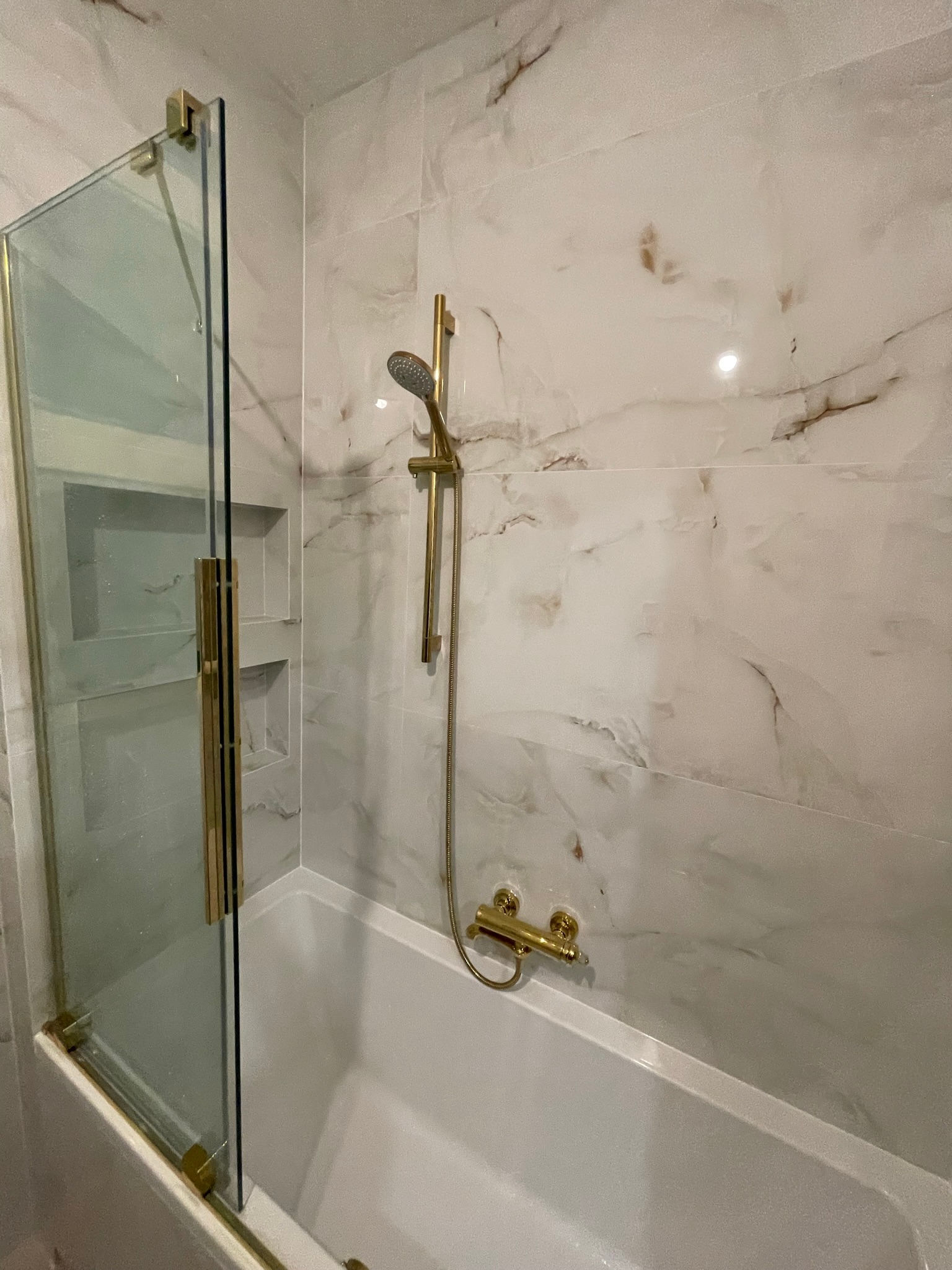 Wykończona łazienka z wanną, marmurowe płytki na ścianach, złota armatura i szklany parawan nawannowy z złotymi detalami.