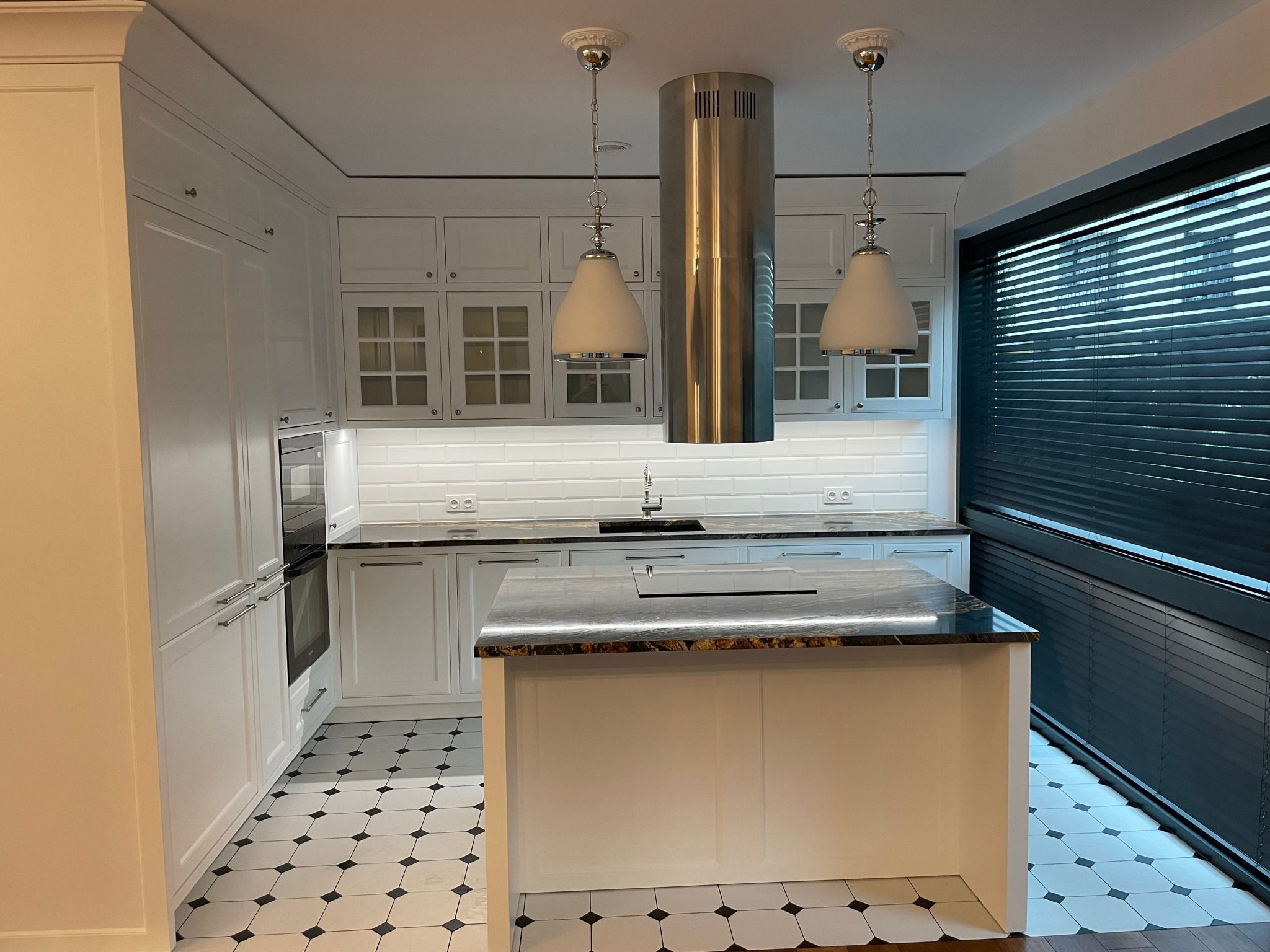 Nowoczesna, jasna kuchnia z wyspą, białymi szafkami z frezowanymi frontami, czarnym blatem z marmurkowym wzorem, płytkami podłogowymi w biało-czarną szachownicę i cylindrycznym okapem ze stali...