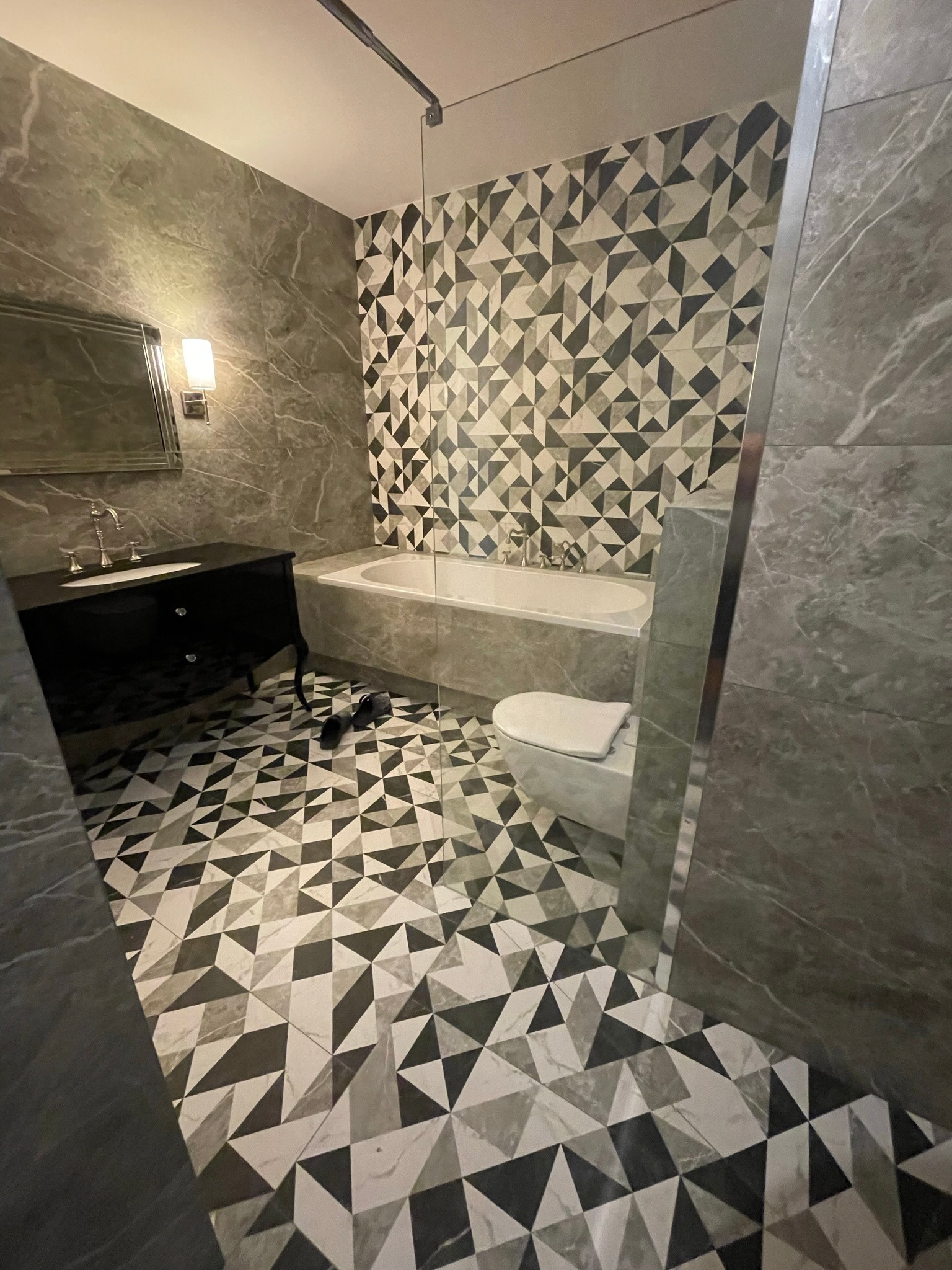 Elegancka łazienka w odcieniach szarości z geometrycznym wzorem na podłodze i ścianie za wanną, czarna szafka pod umywalką w stylu retro, widoczne kapcie na podłodze.
