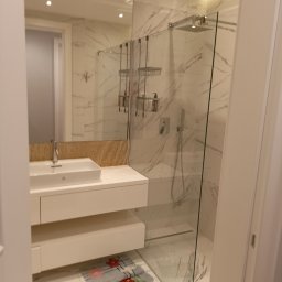 Usługi remontowo budowlano stolarskie - Nowoczesna łazienka z marmurowymi ścianami, szklanym prysznicem typu walk-in, białą szafką z umywalką i mozaiką nad nią, oraz dywanikiem pod szafką.