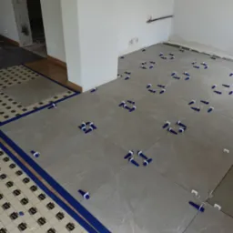 Układanie jasnych płytek podłogowych z użyciem niebieskich krzyżyków dystansowych w nowym, jasnym pomieszczeniu. Widoczna mata podkładowa.