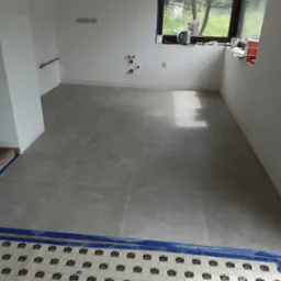 Jasnoszare płytki podłogowe ułożone w kwadraty w pustym pomieszczeniu, częściowo zakryte folią ochronną. Widoczne instalacje na ścianie i okno z widokiem na zieleń.