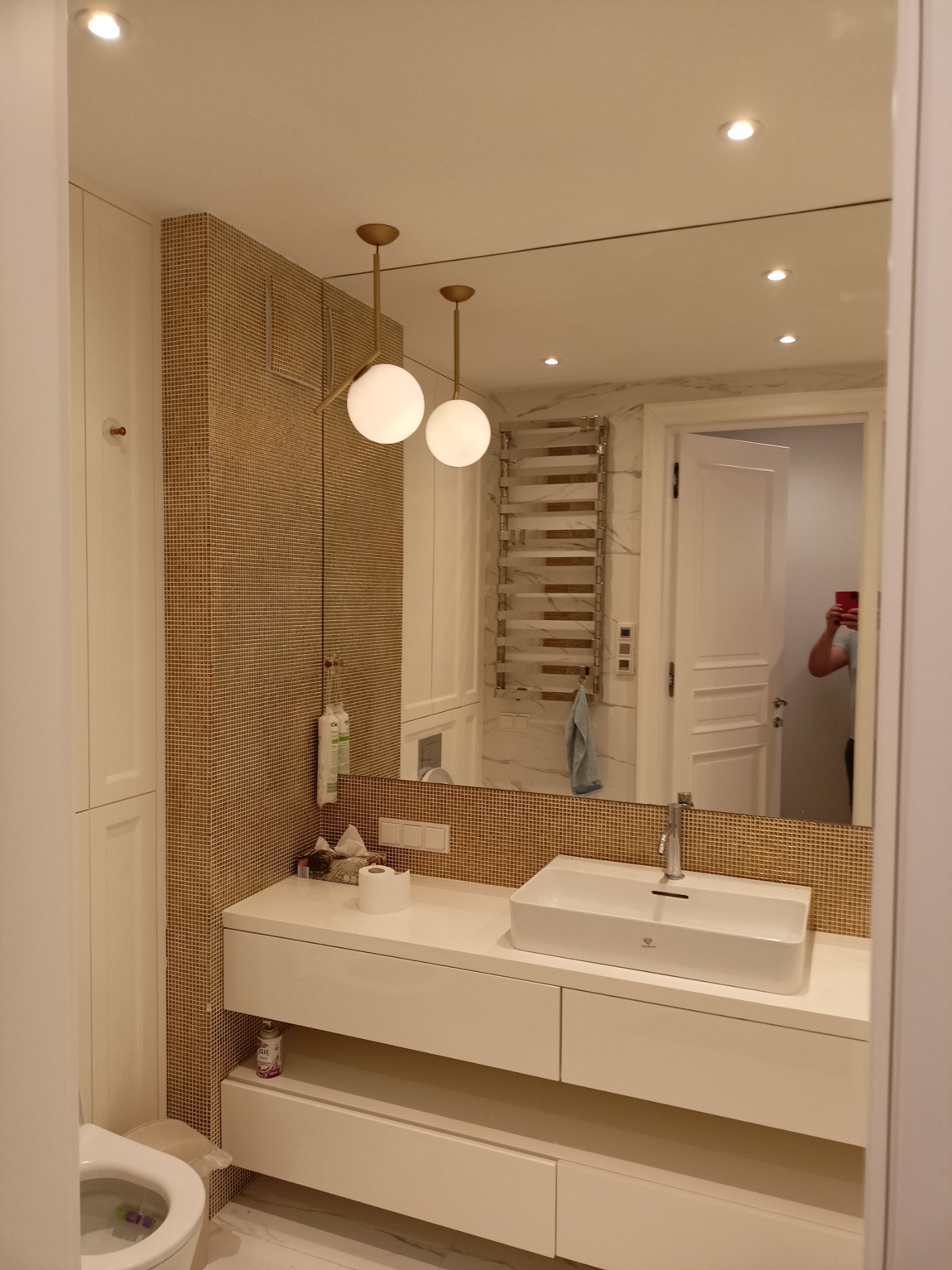 Nowoczesna łazienka z umywalką nablatową, szafką pod umywalką z szufladami, mozaiką w odcieniach złota na ścianie, okrągłymi lampami wiszącymi i grzejnikiem drabinkowym, widok w lustrze na otwarte...