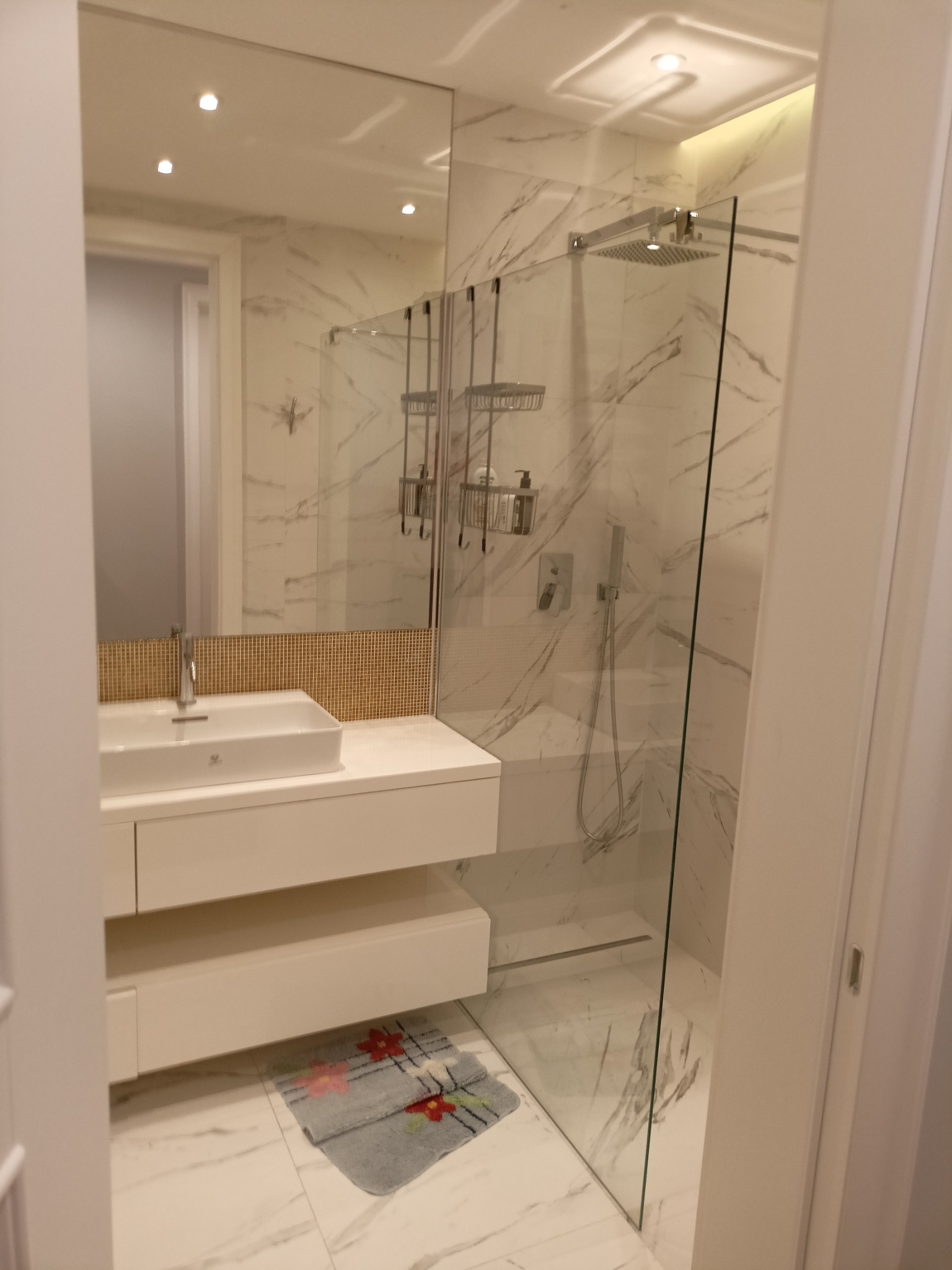 Nowoczesna łazienka z marmurowymi ścianami, szklanym prysznicem typu walk-in, białą szafką z umywalką i mozaiką nad nią, oraz dywanikiem pod szafką.