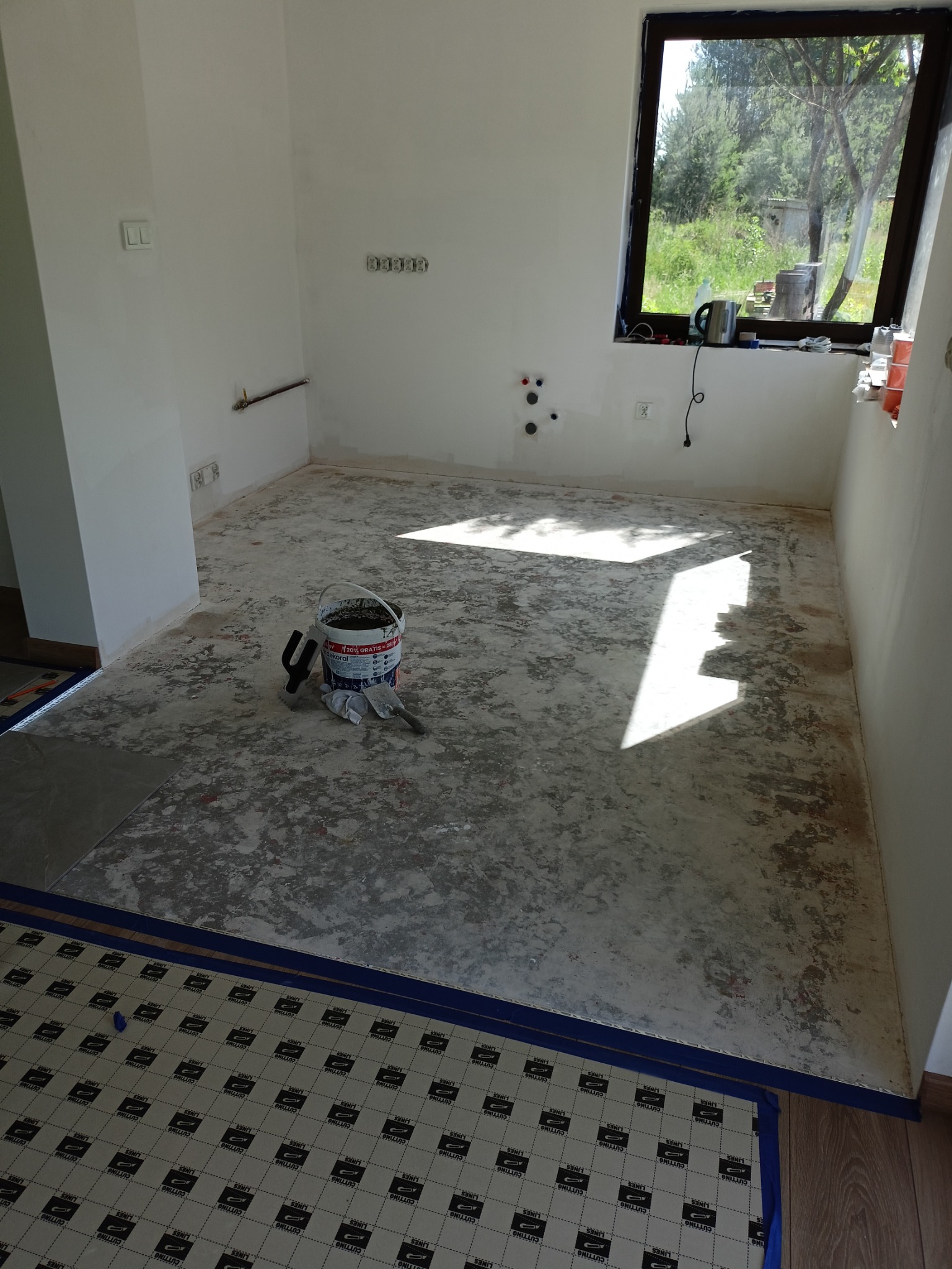 Przygotowanie do układania paneli w jasnym pokoju z oknem. Na podłodze folia ochronna, wiadro z zaprawą i narzędzia. Widoczne niedoskonałości ścian.