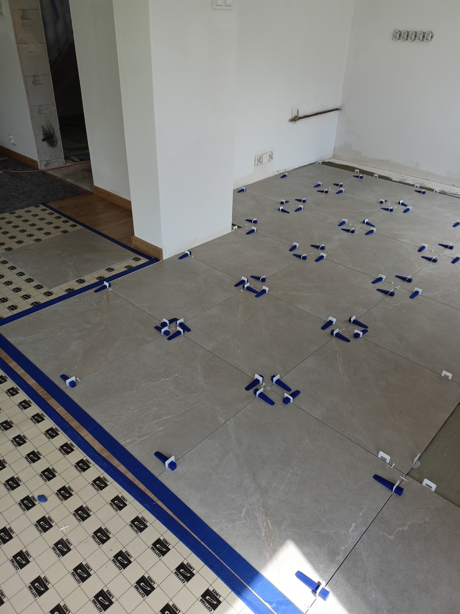 Układanie jasnych płytek podłogowych z użyciem niebieskich krzyżyków dystansowych w nowym, jasnym pomieszczeniu. Widoczna mata podkładowa.