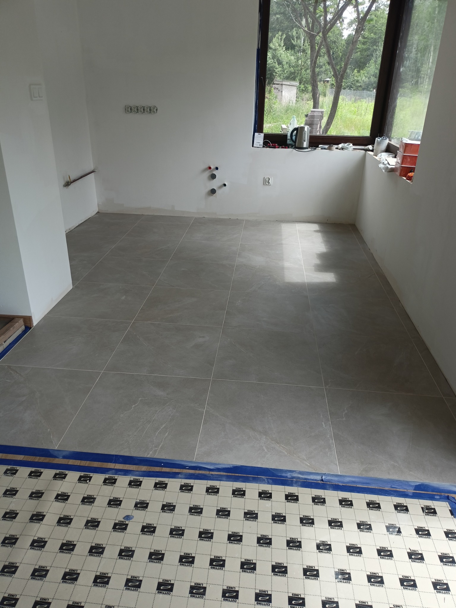 Jasnoszare płytki podłogowe ułożone w kwadraty w pustym pomieszczeniu, częściowo zakryte folią ochronną. Widoczne instalacje na ścianie i okno z widokiem na zieleń.