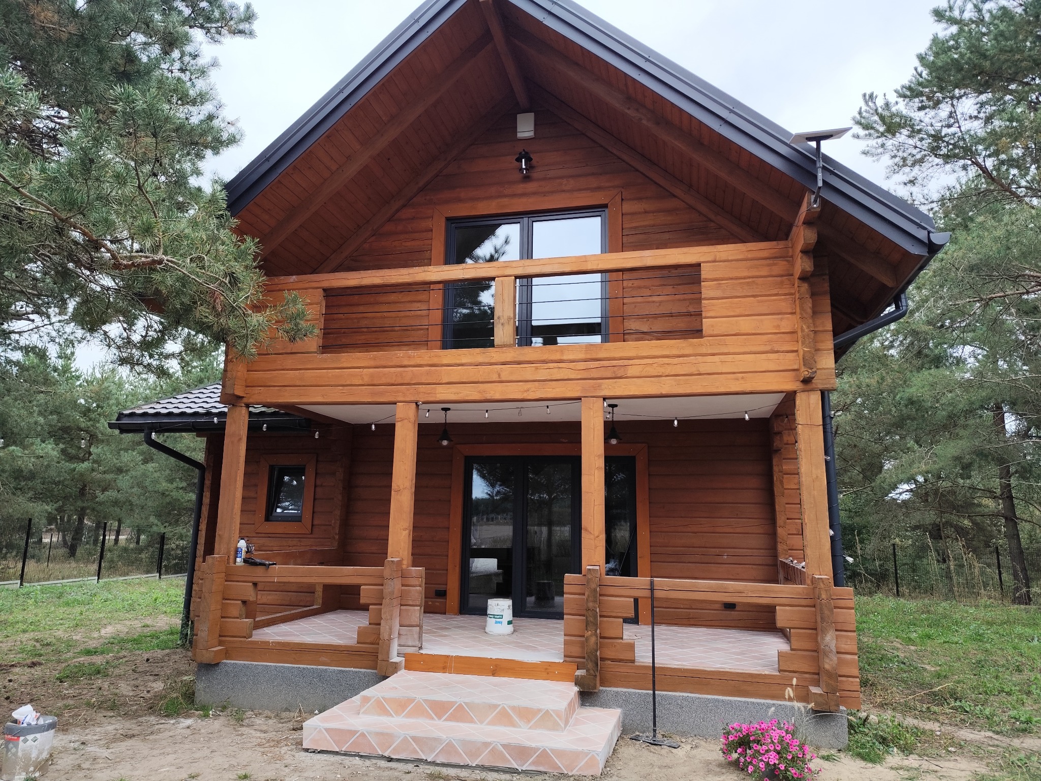 Drewniany dom z balkonem i tarasem, wykończony w naturalnym stylu, otoczony zielenią. Widoczne detale wykończenia i oświetlenie zewnętrzne.