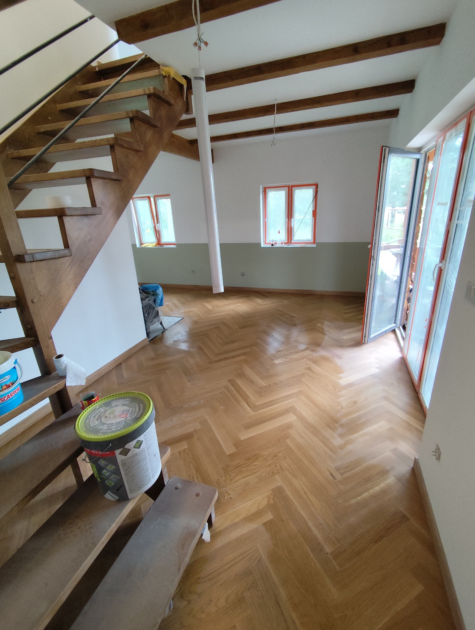 Jasny parkiet ułożony w jodełkę w przestronnym wnętrzu z drewnianymi schodami i belkami stropowymi. Widoczne okna i drzwi balkonowe. Wiadro farby na schodach.