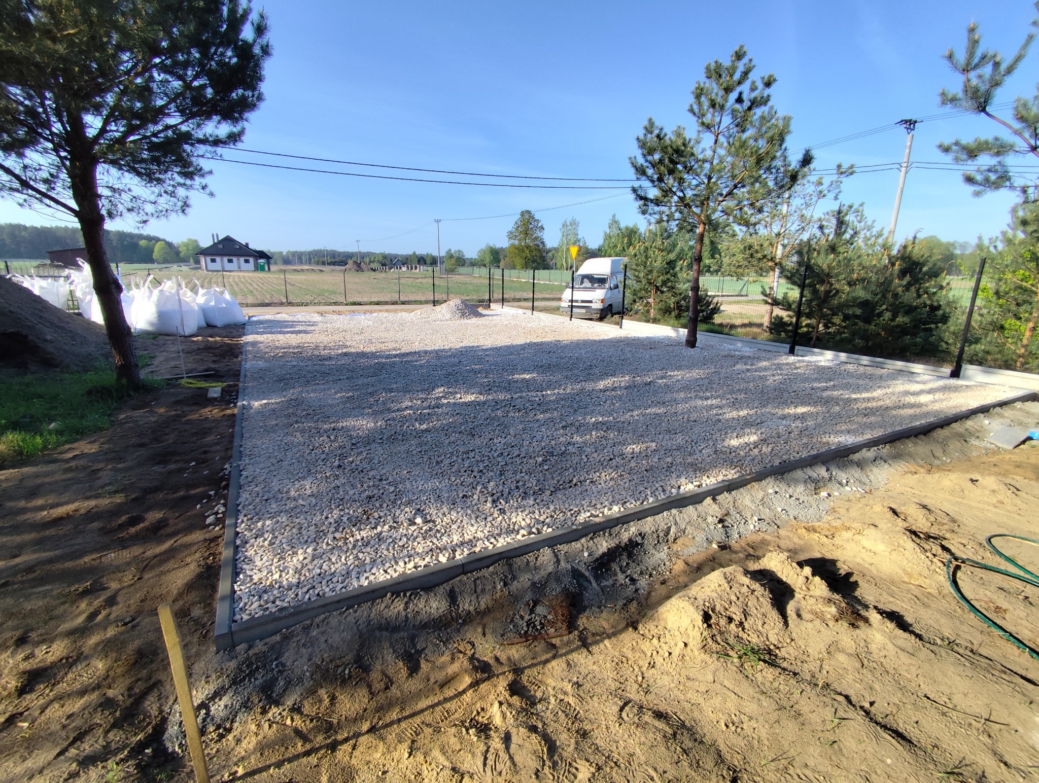 Przygotowany teren pod budowę: wyrównany, wysypany żwirem, obrzeża z betonowych krawężników. W tle białe worki z materiałem budowlanym i zaparkowany biały bus.