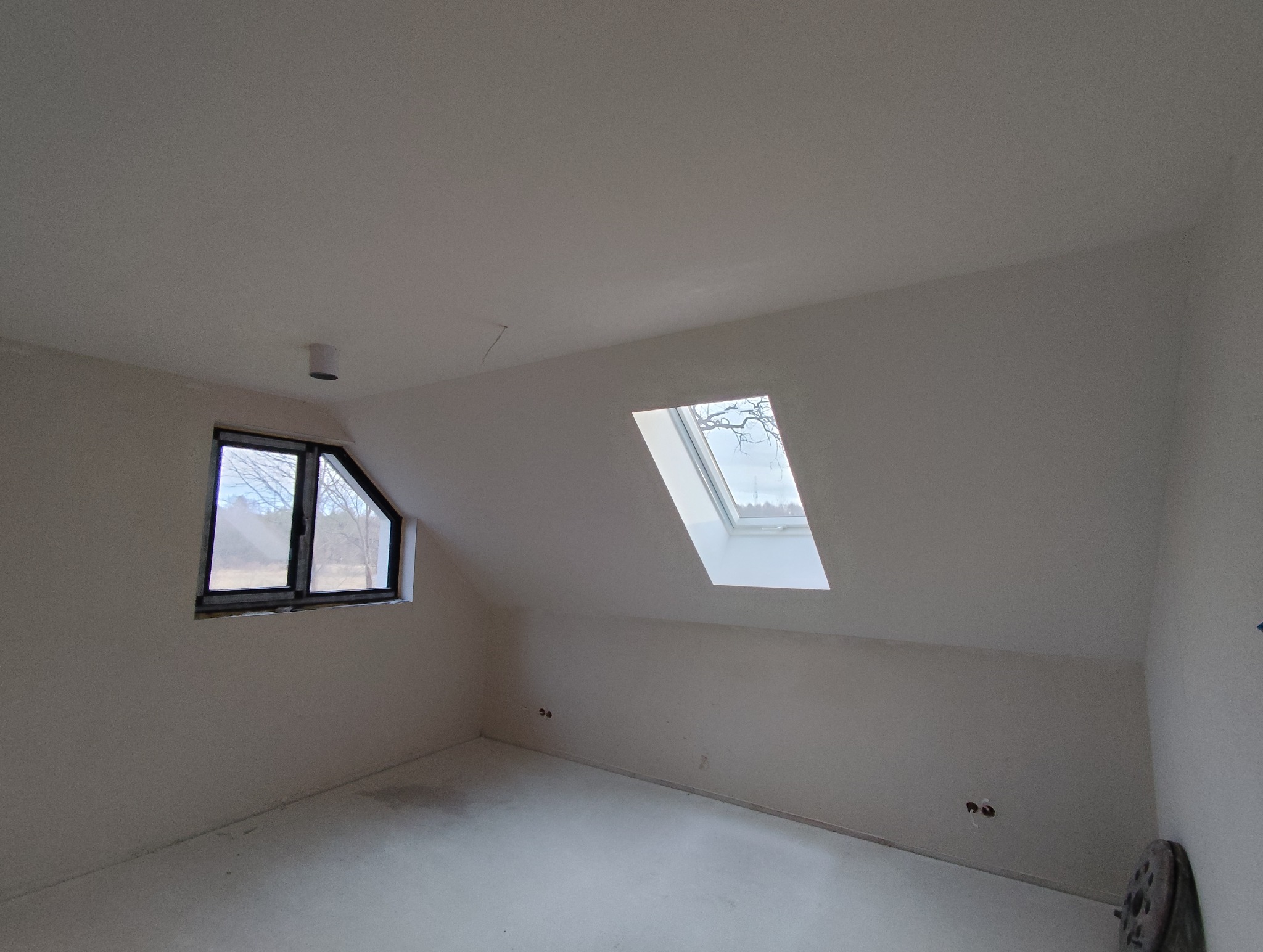 Poddasze po szpachlowaniu: gładkie, białe ściany i sufit, okno dachowe i zwykłe okno. Widoczne ślady po gniazdkach elektrycznych. Przy podłodze fragment szlifierki.