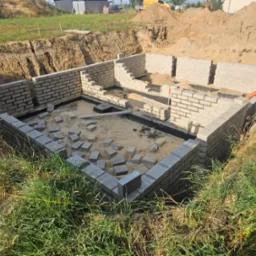 Wykop pod fundamenty domu murowanego z częściowo wzniesionymi ścianami z bloczków betonowych, widoczne niezwiązane bloczki wewnątrz fundamentu, pomarańczowa rura w tle, wykopana ziemia i trawa wokół.