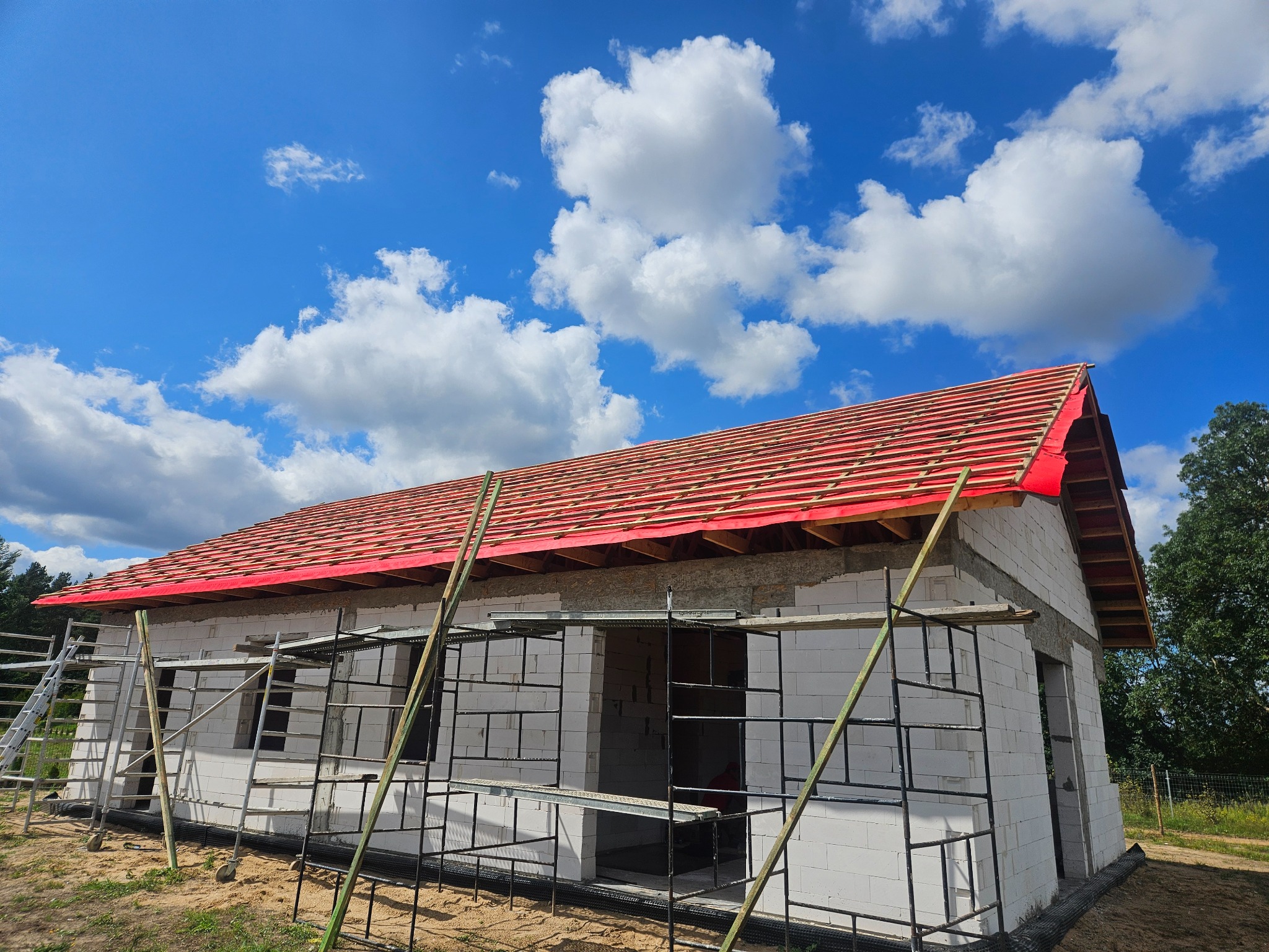 Budowa domu z betonu komórkowego w trakcie wznoszenia ścian i konstrukcji dachu, widoczne rusztowania i czerwona folia dachowa pod pokrycie, na tle błękitnego nieba z białymi chmurami.