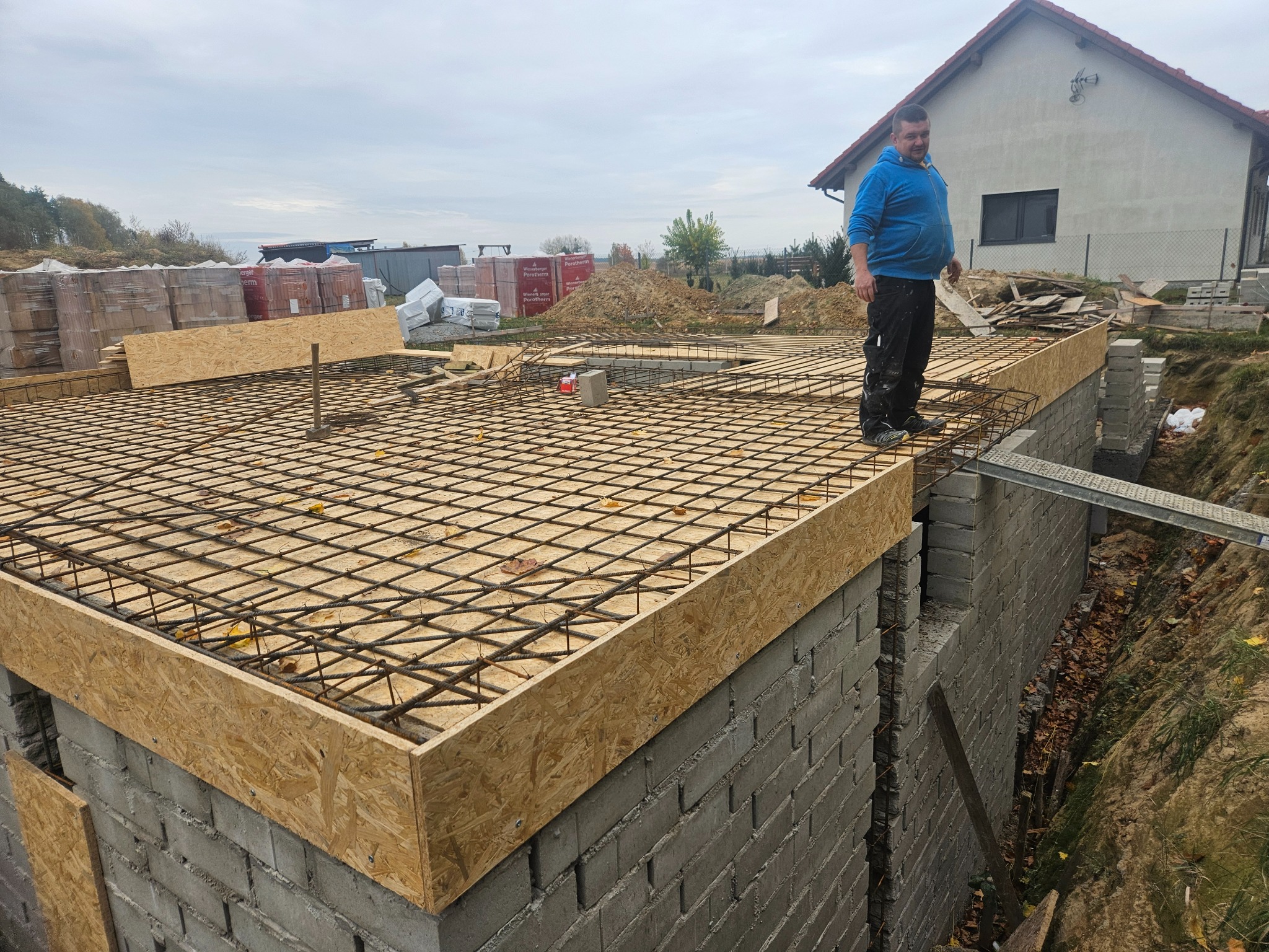 Fundament domu w trakcie budowy: zbrojenie stalowe, szalunek z płyty OSB, bloczki betonowe, pracownik w niebieskiej bluzie stojący na konstrukcji, w tle dom jednorodzinny w stanie surowym otwartym.
