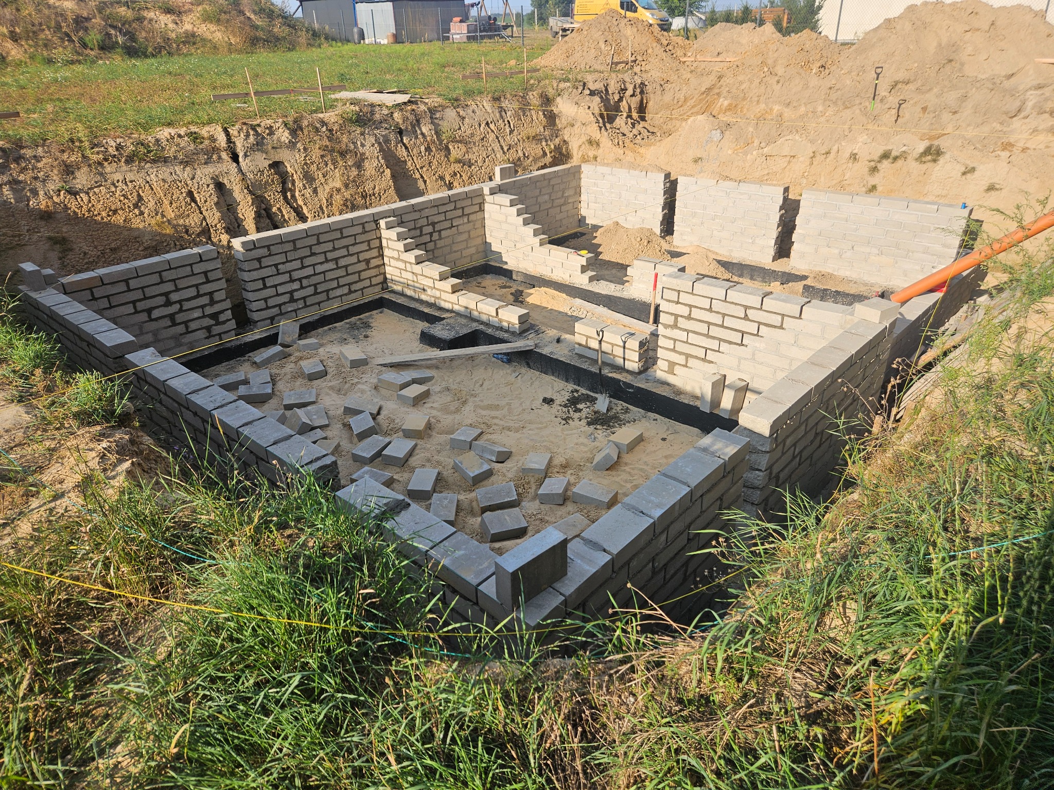 Wykop pod fundamenty domu murowanego z częściowo wzniesionymi ścianami z bloczków betonowych, widoczne niezwiązane bloczki wewnątrz fundamentu, pomarańczowa rura w tle, wykopana ziemia i trawa wokół.