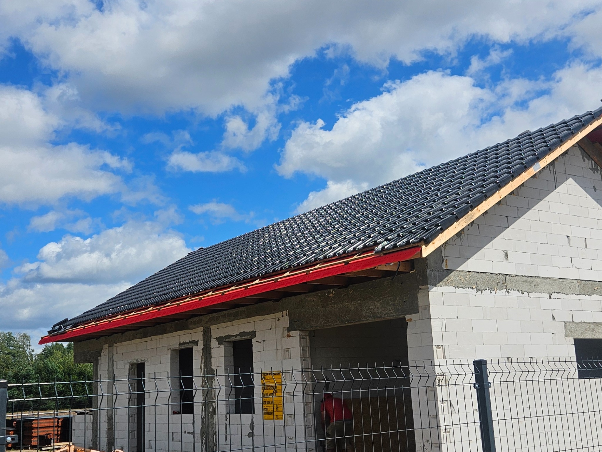 Budowa domu z białych bloczków, widoczny fragment dachu pokrytego czarną dachówką, czerwona listwa okapowa, błękitne niebo w tle.