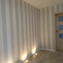 Pionowe, szaro-białe pasy tapety na ścianie z oświetleniem podłogowym i fragmentem drzwi z przeszkleniami. Neutralna kolorystyka wnętrza.