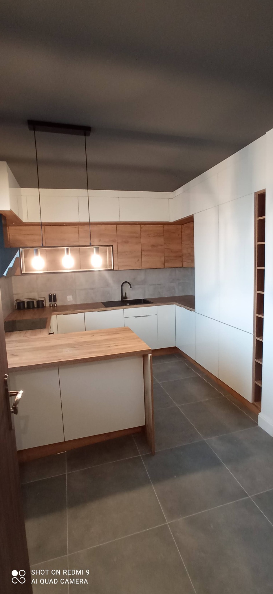 Nowoczesna kuchnia z białymi szafkami, drewnianymi akcentami i wyspą, szarymi płytkami na ścianie i podłodze oraz podwieszanymi lampami z żarówkami edisona.