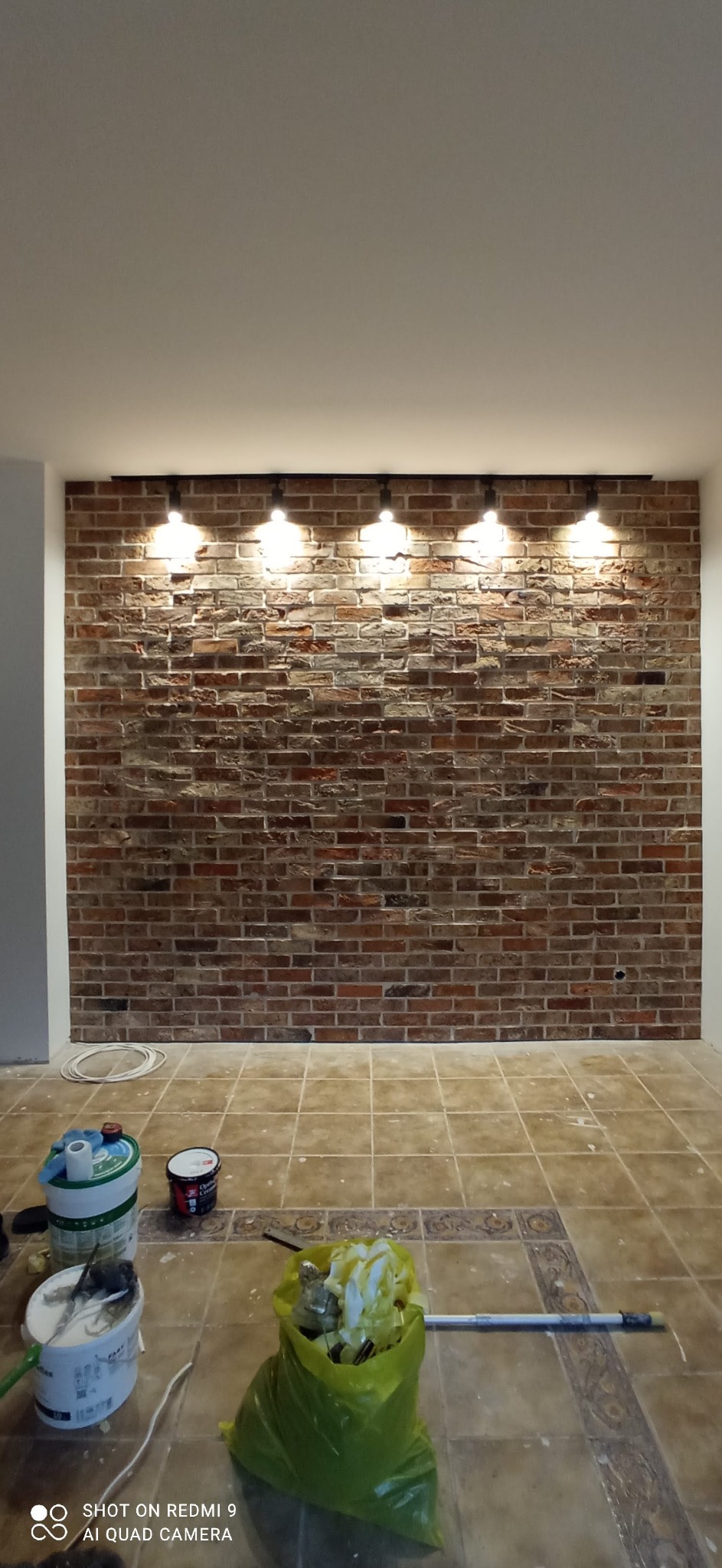 Ściana z cegieł dekoracyjnych z czterema punktowymi lampami, widoczne puszki farb i worek na śmieci na podłodze z płytek.