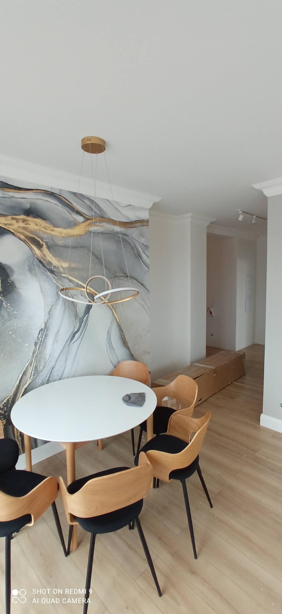 Nowoczesne wnętrze jadalni z okrągłym białym stołem, krzesłami z drewnianymi oparciami i czarnymi siedziskami, designerską lampą wiszącą oraz tapetą w odcieniach szarości i złota, świeżo ułożona...