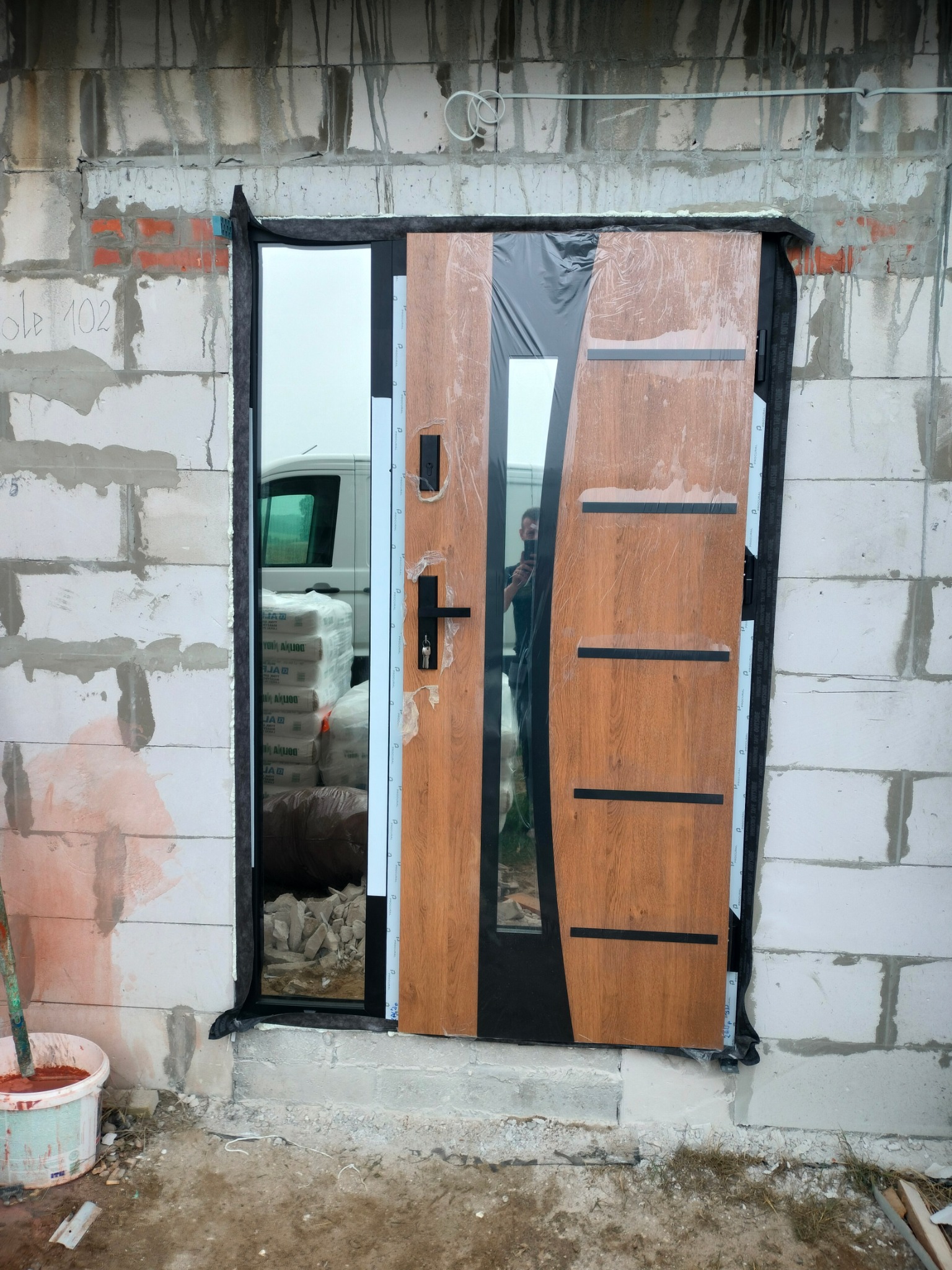 Nowe drzwi wejściowe z szybą, częściowo zabezpieczone folią, zamontowane w surowym murze z betonu komórkowego. Widoczny kubeł z zaprawą i worki w tle.