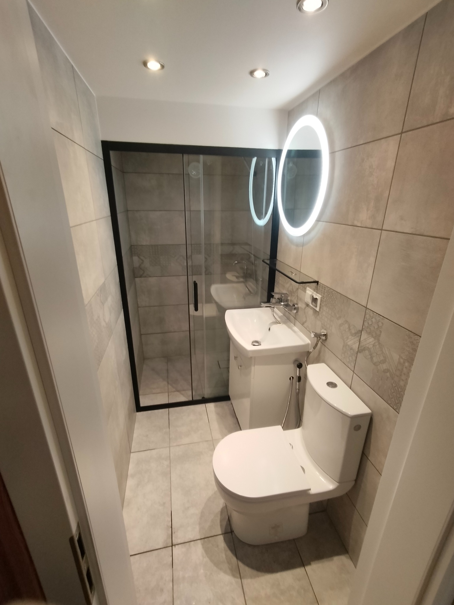 Nowoczesna łazienka po remoncie: kabina prysznicowa z czarną ramą, umywalka z szafką, podświetlane lustro i toaleta. Płytki w odcieniach szarości z wzorami.