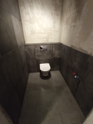 Nowoczesna toaleta w Dobrczu po remoncie. Szare płytki na ścianach do połowy wysokości, biała miska WC, surowe ściany powyżej. Minimalistyczny design.