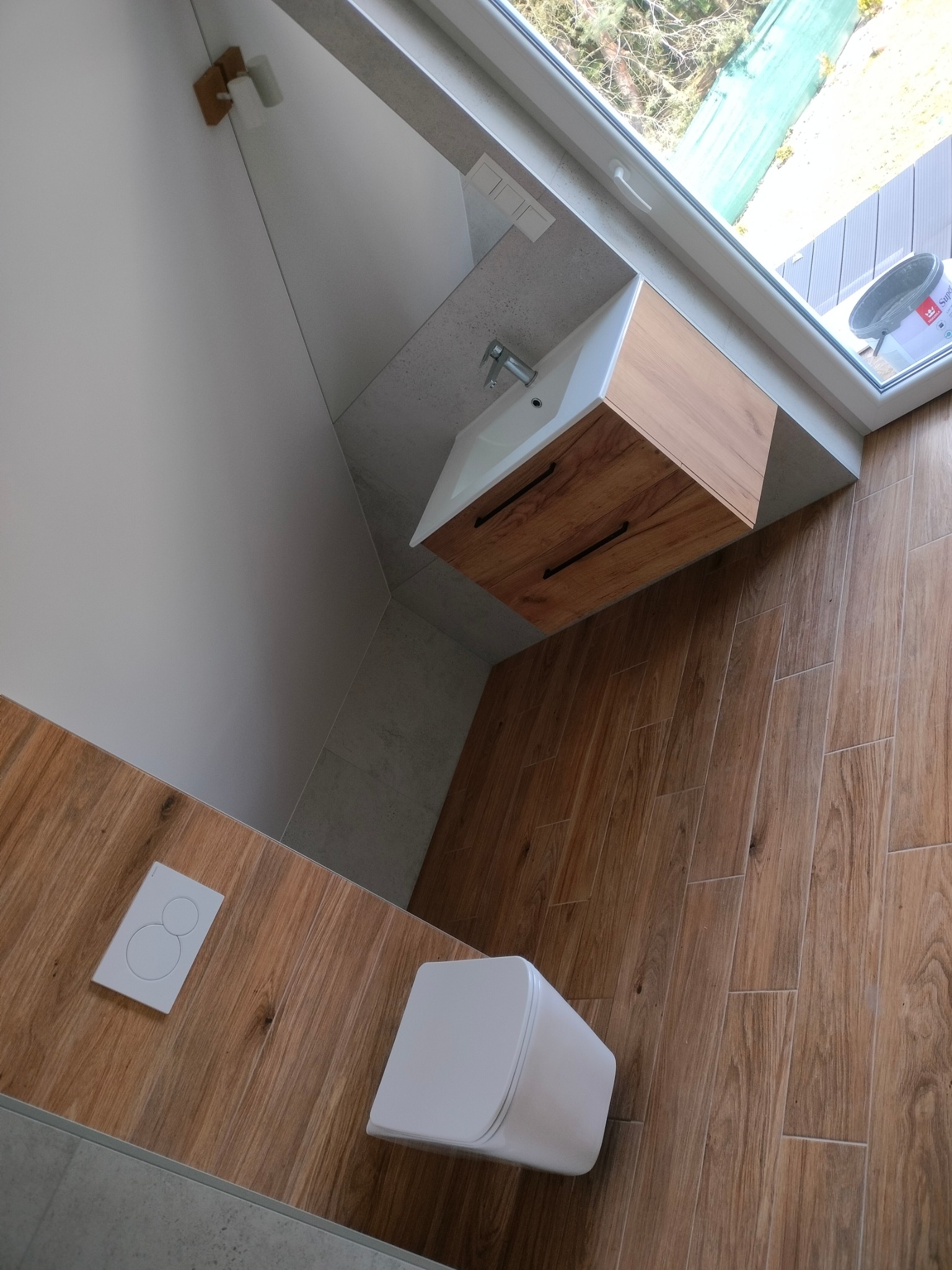 Drewnopodobne płytki podłogowe w łazience z szafką umywalkową i toaletą. Widoczne okno z wiadrem farby i fragment zielonego ogrodzenia za nim.