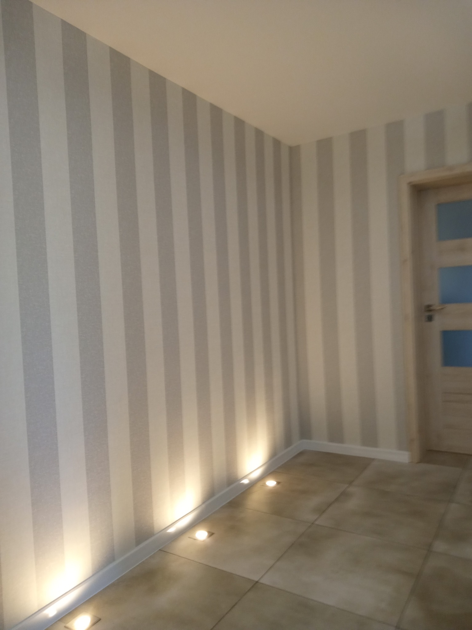 Pionowe, szaro-białe pasy tapety na ścianie z oświetleniem podłogowym i fragmentem drzwi z przeszkleniami. Neutralna kolorystyka wnętrza.