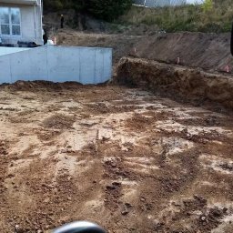 Zbyszek Ko - Wykopany teren z betonową ścianą oporową w tle, widoczne paliki wyznaczające granice, ziemia częściowo wilgotna, odbicie w lusterku pojazdu po prawej stronie.