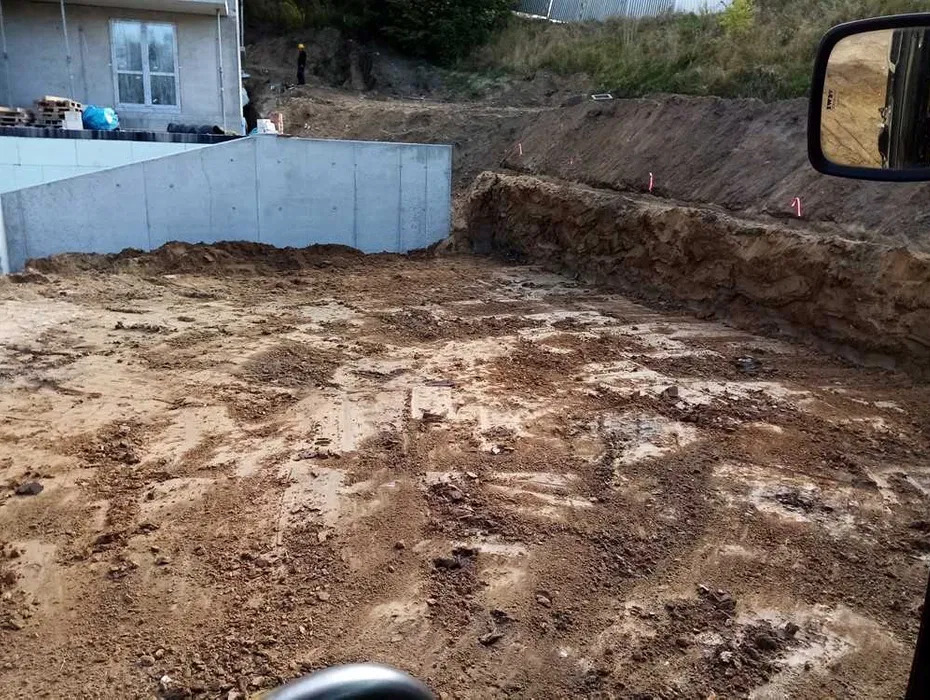 Wykopany teren z betonową ścianą oporową w tle, widoczne paliki wyznaczające granice, ziemia częściowo wilgotna, odbicie w lusterku pojazdu po prawej stronie.