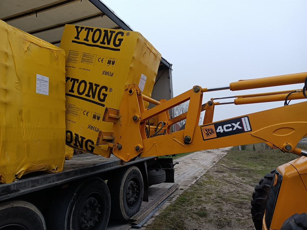 Żółty podnośnik teleskopowy JCB 4CX ładuje palety z bloczkami Ytong na platformę ciężarówki; bloczki owinięte folią ochronną.