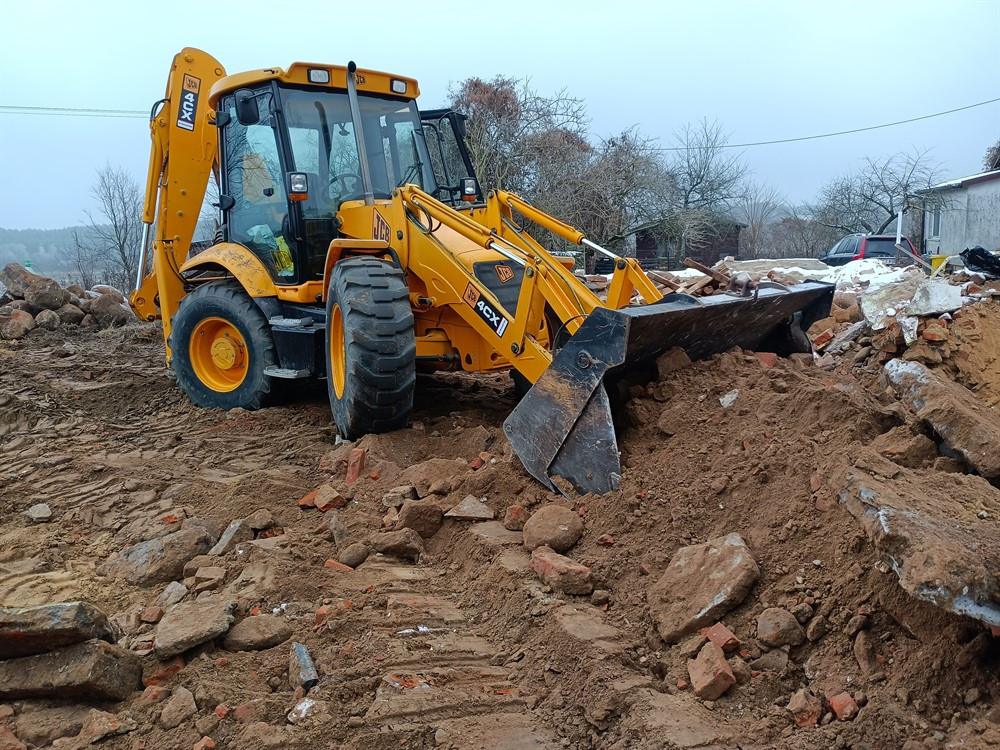 Żółta koparko-ładowarka JCB 4CX na nierównym terenie z gruzem i ziemią, widoczne ślady po pracy maszyny, w tle budynek i drzewa pod pochmurnym niebem.