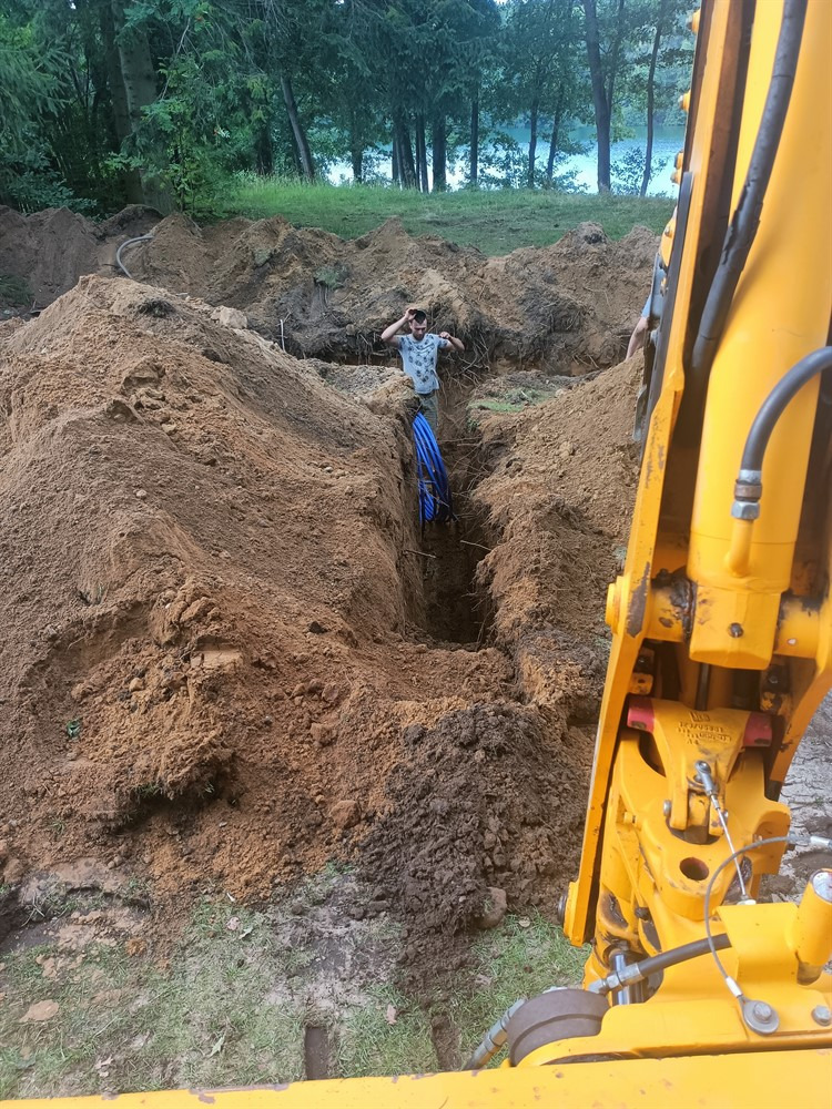 Głęboki wykop w ziemi z ułożonymi niebieskimi rurami, widok z kabiny żółtej koparki, pracownik w dole wykopu z rękami uniesionymi nad głową, w tle drzewa i jezioro.