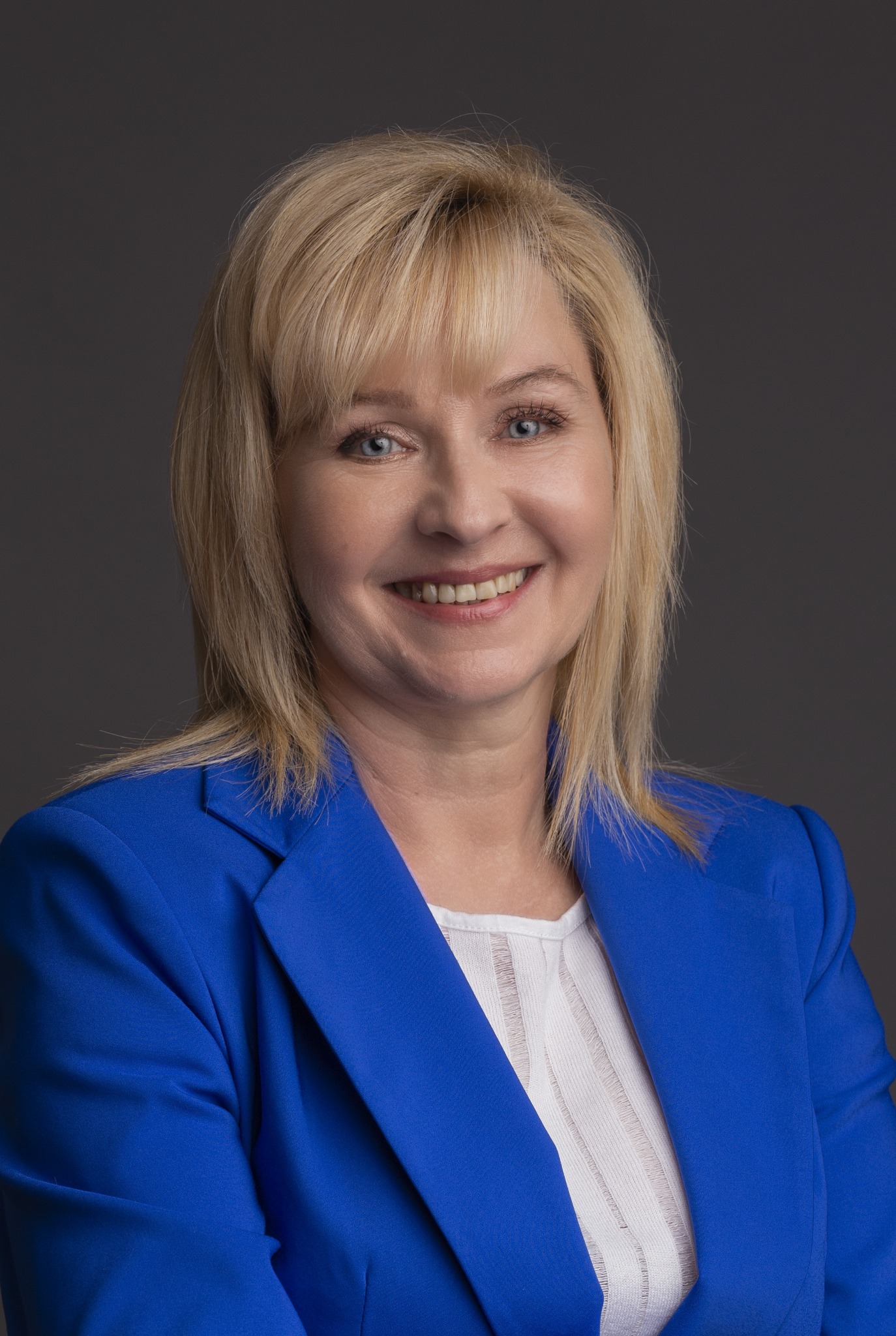 Portret uśmiechniętej kobiety z blond włosami w niebieskim żakiecie na szarym tle.