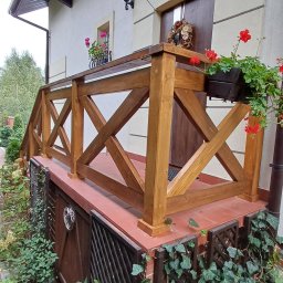 Redwood Tarasy Drewniane - Drewniana balustrada z krzyżowym wzorem na niewielkim tarasie przed domem, z donicami z czerwonymi kwiatami, widok z dołu.