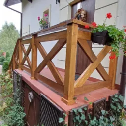 Drewniana balustrada z krzyżowym wzorem na niewielkim tarasie przed domem, z donicami z czerwonymi kwiatami, widok z dołu.