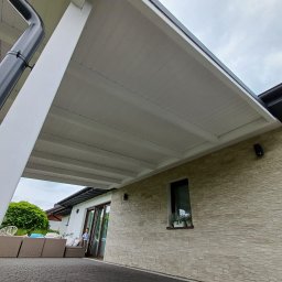 Redwood Tarasy Drewniane - Zadaszona przestrzeń tarasowa z białym sufitem, widok z dołu, z widocznym fragmentem elewacji z kamienia dekoracyjnego i meblami ogrodowymi w tle.