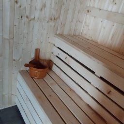 Redwood Tarasy Drewniane - Wnętrze sauny wykończone jasnym drewnem, z drewnianymi ławami i cebrzykiem z chochlą na jednej z nich.